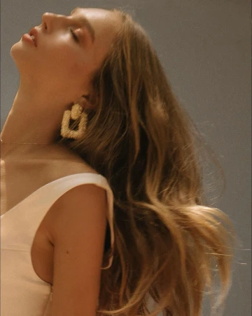Blonde woman in a sleeveless white top, poses indoors with eyes closed. She wears statement square earrings, with grey backdrop.