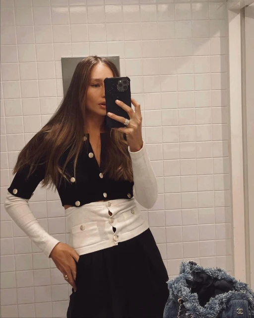 Woman takes mirror selfie wearing a black-and-white button-down sweater with black bottoms and long brown hair in an indoor setting.