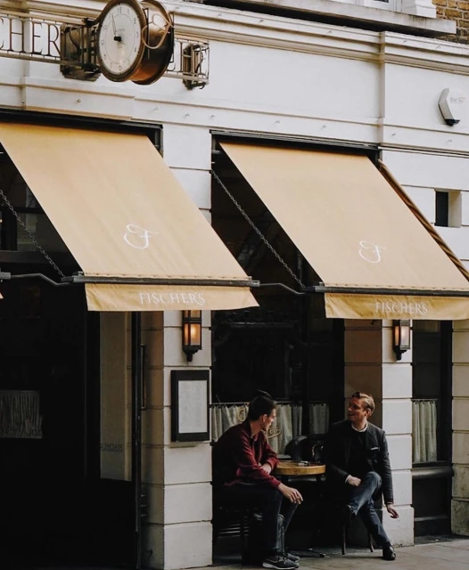 Two people sitting at a cafe table outside Fischer's restaurant, under beige awnings, with a vintage clock on the building's facade.