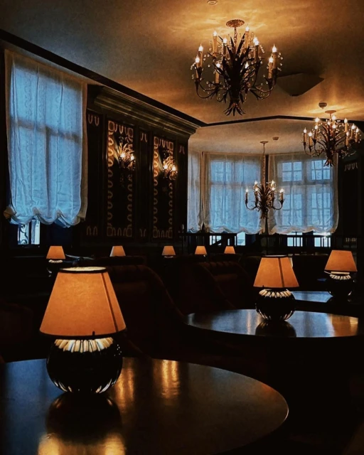 Warmly lit interior of a lounge area featuring round tables with lamps, soft seating, decorative wall panels, and elegant chandeliers.