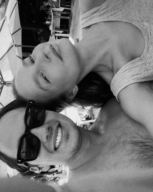 Black and white photo of a smiling man in sunglasses and a woman lying together, possibly outdoors, showing a close and personal moment.