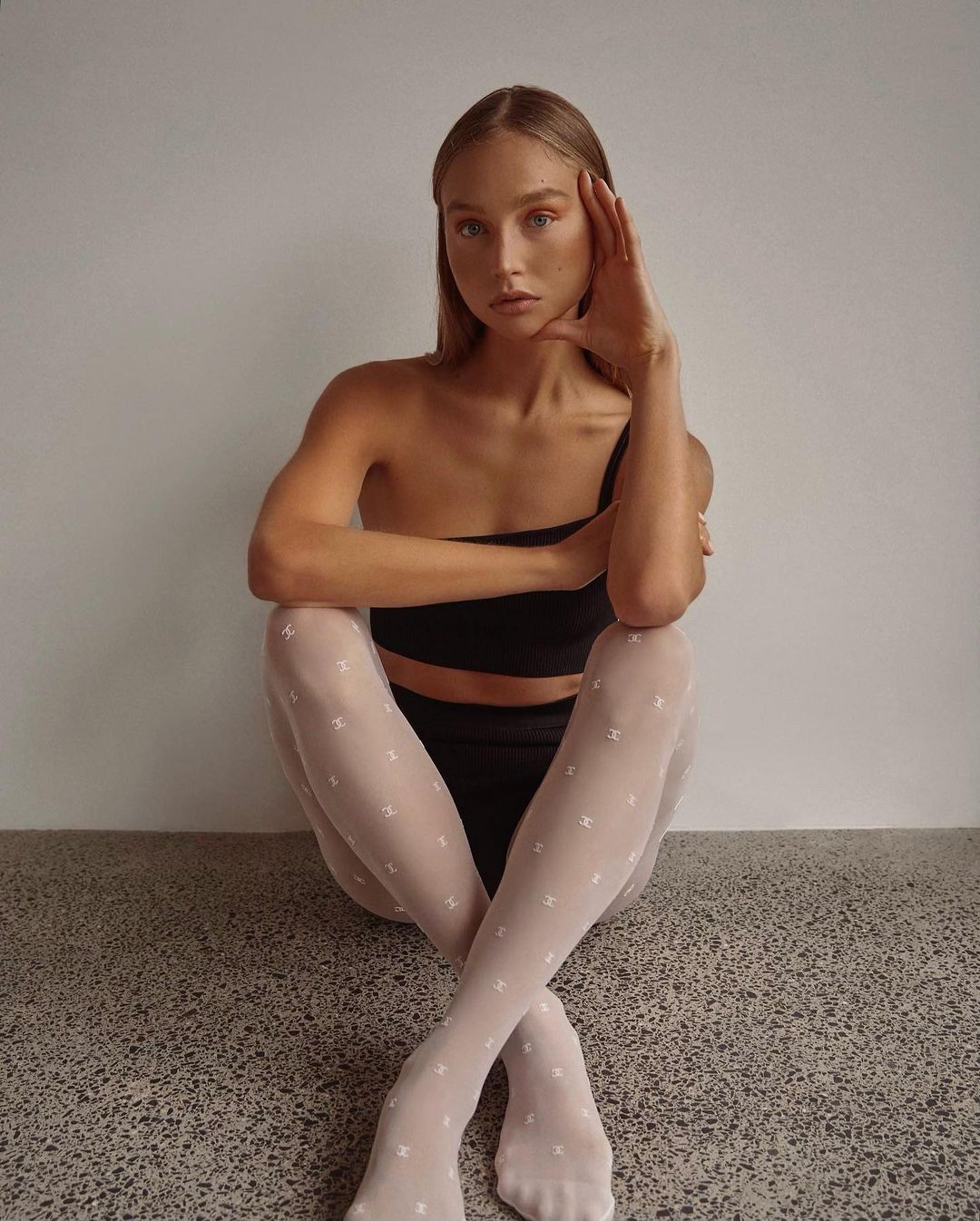 Woman in black top and white patterned tights sitting indoors against a light wall with a simple gray floor.