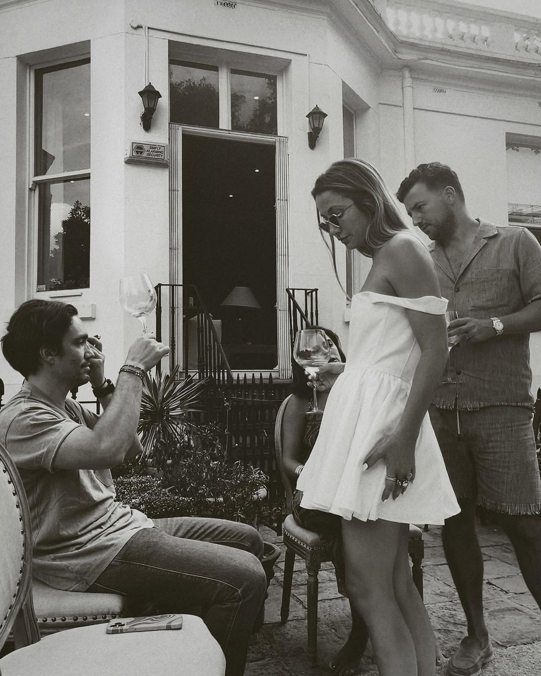 Outdoor social gathering on a patio with a man raising a wine glass, a woman in a short dress, and another man standing nearby.