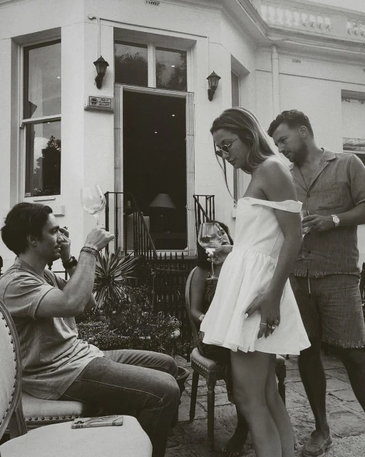 Outdoor social gathering on a patio with a man raising a wine glass, a woman in a short dress, and another man standing nearby.
