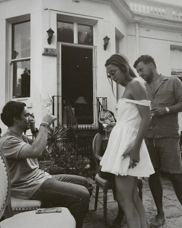 Outdoor social gathering on a patio with a man raising a wine glass, a woman in a short dress, and another man standing nearby.