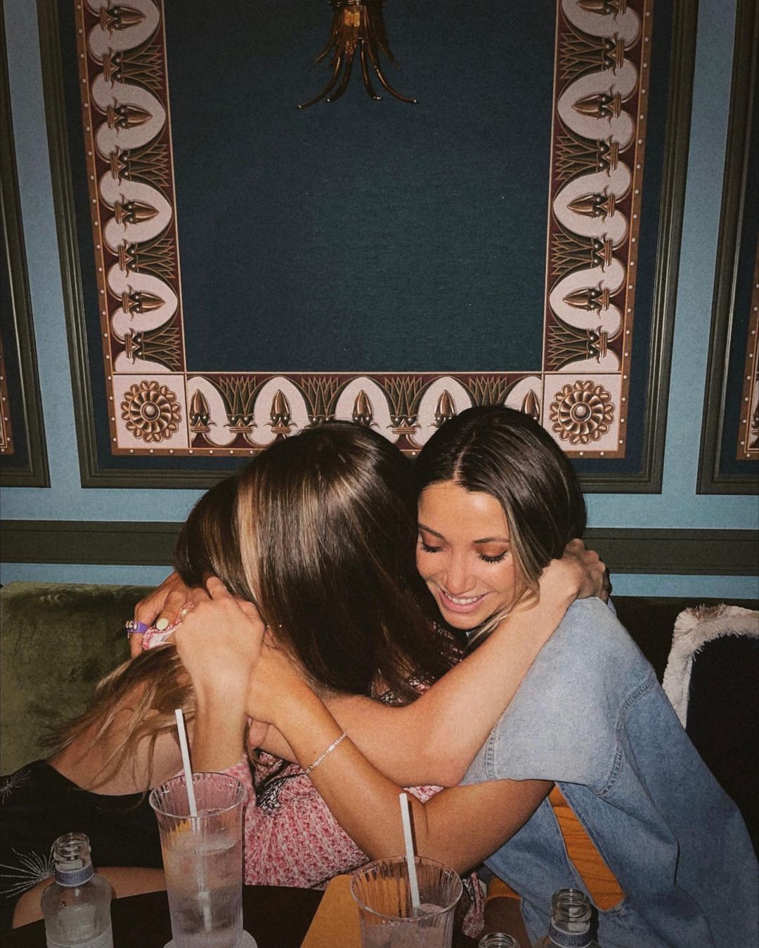 Three young adult women embrace indoors at a table with beverages in glasses. Decorative wall art is visible in the background.
