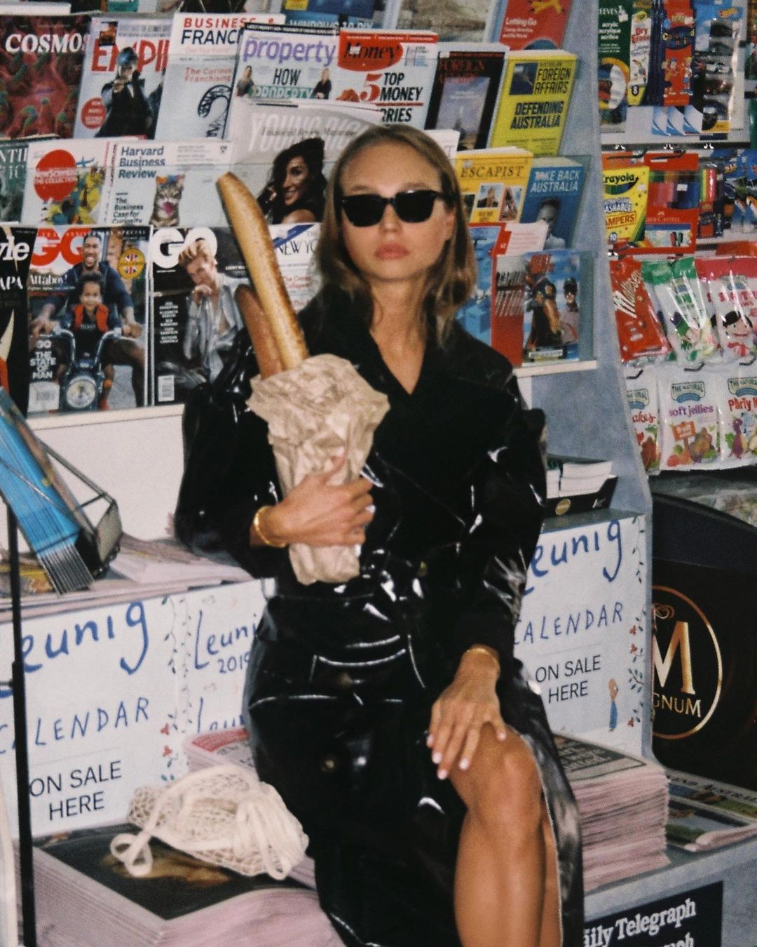Woman in a black vinyl trench coat poses with baguettes in front of a magazine rack, wearing sunglasses with hair down, holding a tote bag.