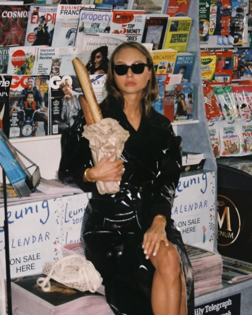 Woman in a black vinyl trench coat poses with baguettes in front of a magazine rack, wearing sunglasses with hair down, holding a tote bag.