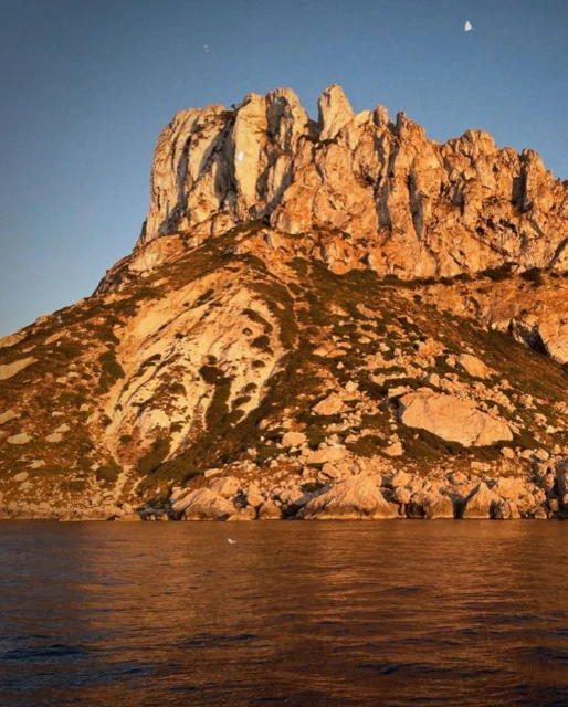 Coastal mountain landscape with golden sunlight illuminating the rocky peak and lush vegetation, set against a deep blue sky and ocean.