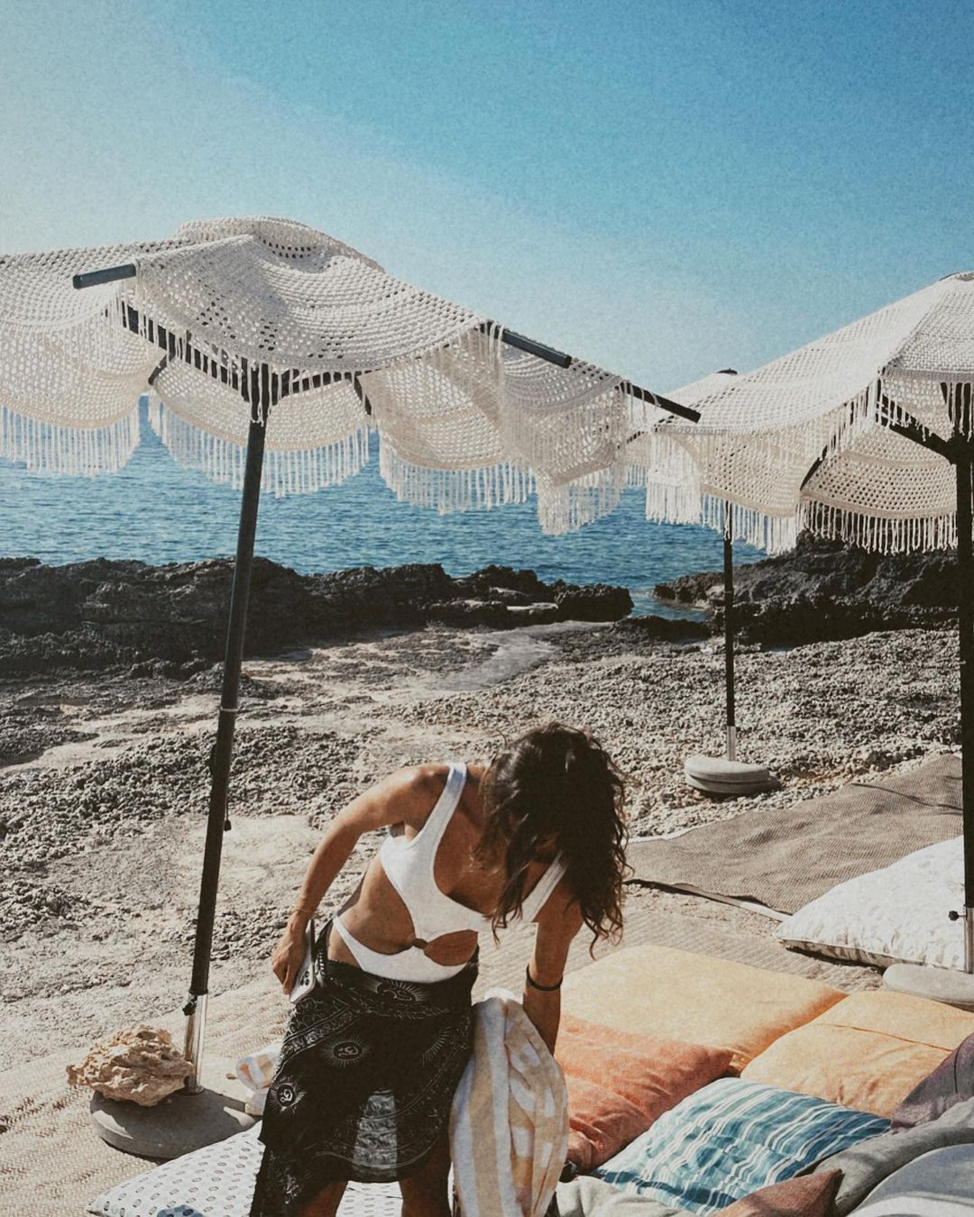 Woman in a white swimsuit and a sarong arranging cushions on a sandy beach under a sun umbrella with ocean and rocky shore backdrop.