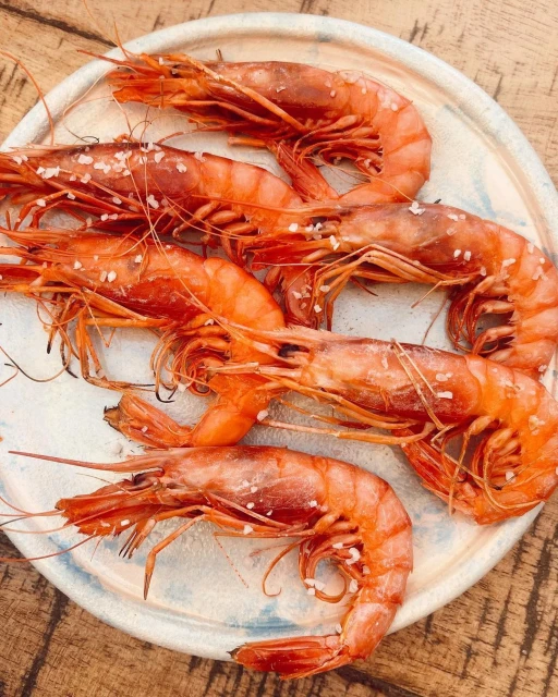 Overhead view of a plate of grilled scarlet shrimp seasoned with coarse sea salt, arranged on a light blue decorative plate atop a wooden surface.