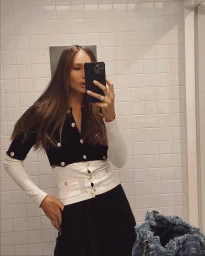 Woman taking mirror selfie in white tiled bathroom wearing black cardigan with gold buttons and white sleeves