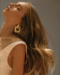 Profile portrait of a woman with long wavy brown hair wearing statement textured earrings and a white top against a grey wall