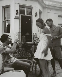 Group of people gathering outside a white building with a woman in a white off-shoulder dress holding a wine glass.