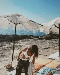 Woman in a white cutout swimsuit and dark patterned sarong standing on a sandy beach beneath crochet fringe beach umbrellas.