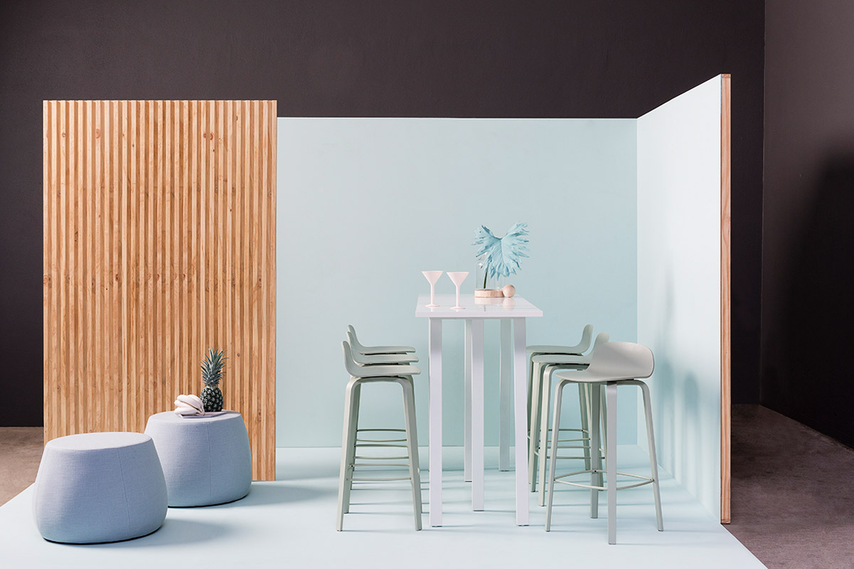Photo in a studio setting showcasing a high table with stools, two poufs, and a pineapple against a background with blue and brown walls.