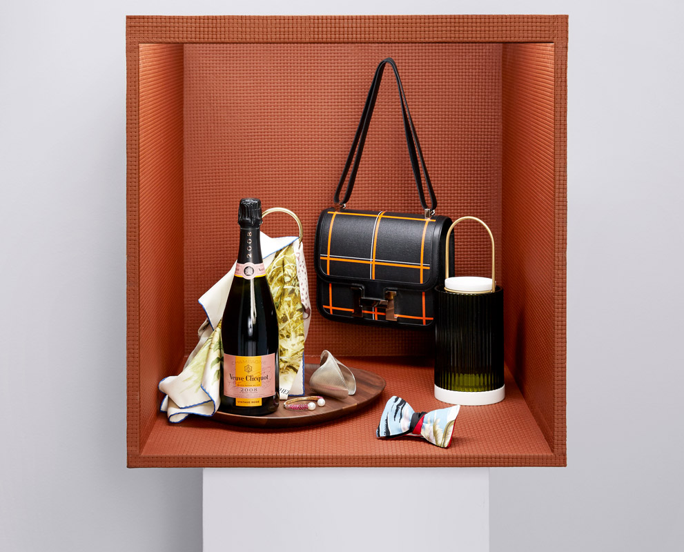 Still life of champagne, handbag, and decorative objects in a recessed orange niche with a white base, studio lighting, product photography.
