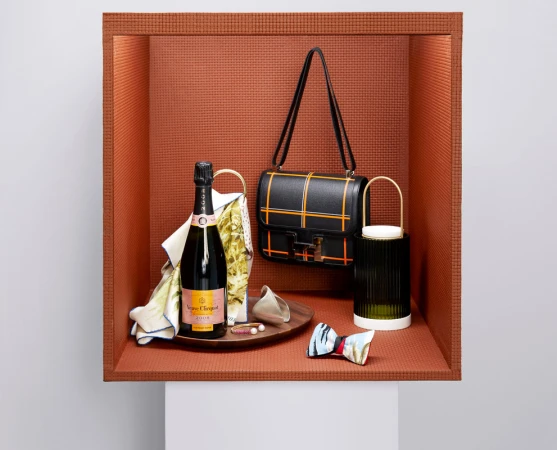 Still life of champagne, handbag, and decorative objects in a recessed orange niche with a white base, studio lighting, product photography.