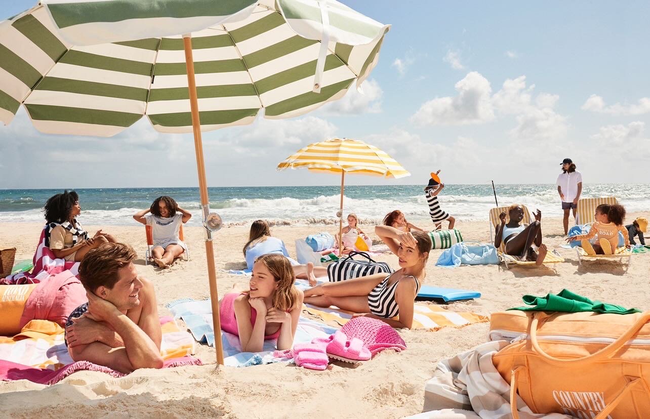 A group of people relax on a sandy beach under umbrellas with ocean waves and blue sky in the background during a sunny day.