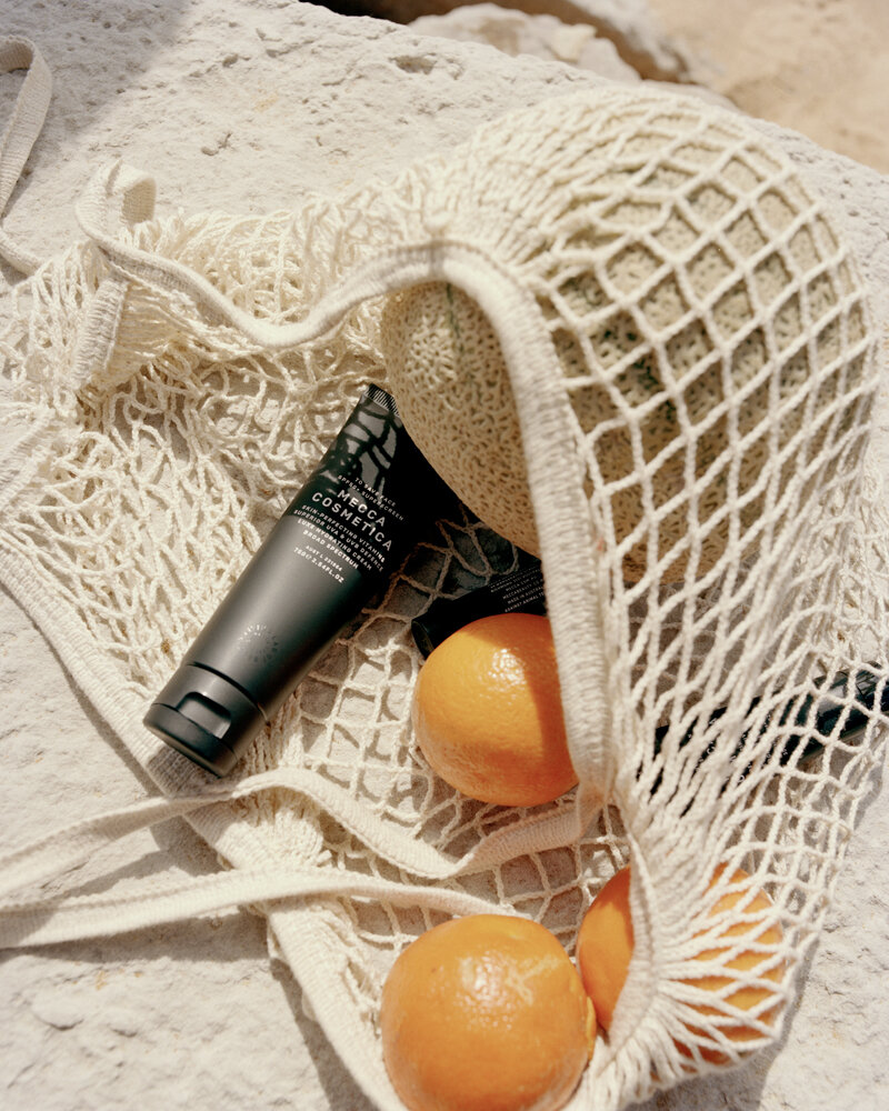 Still life of oranges, a melon, and skincare in a woven bag, set on an outdoor stone surface with soft natural light.