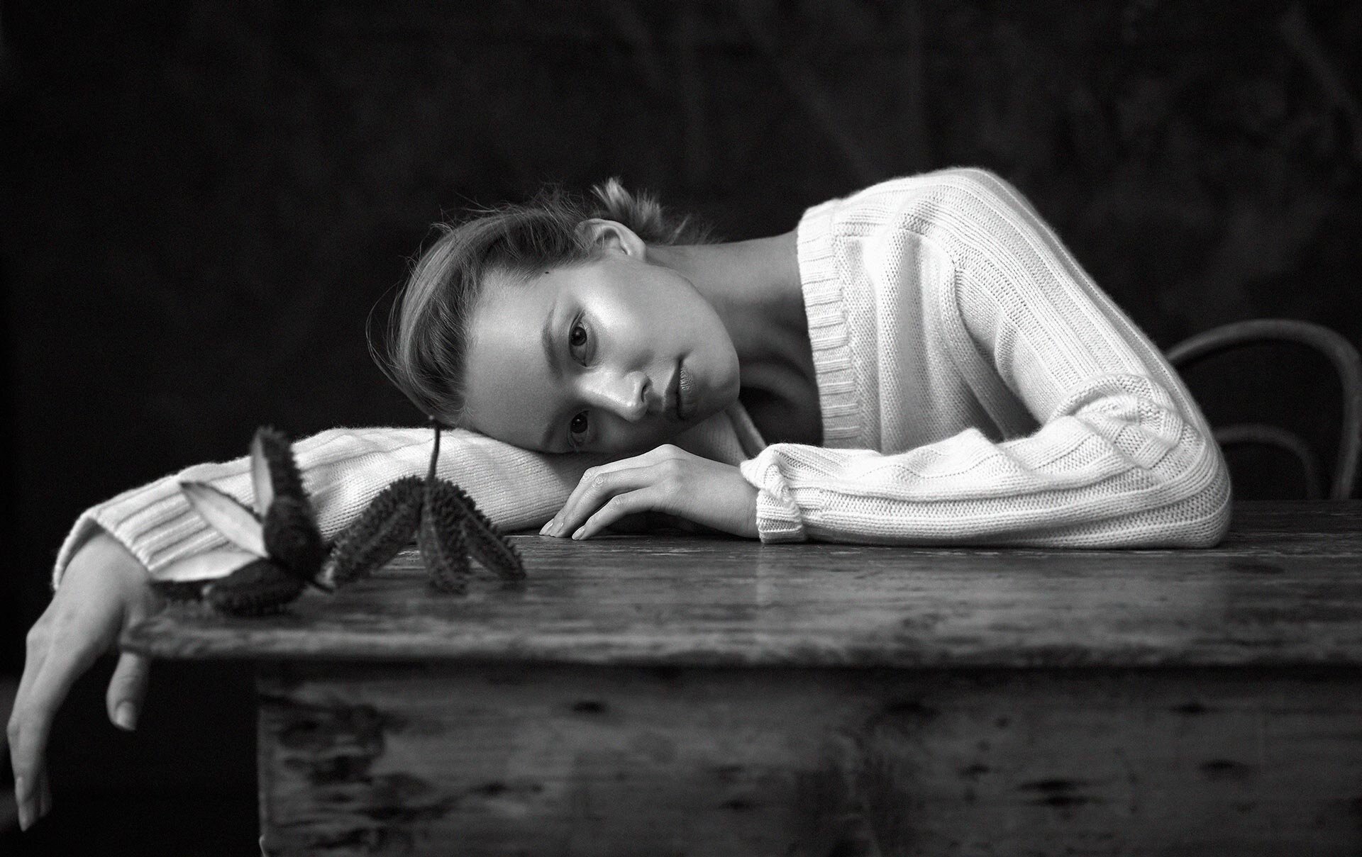 Monochrome portrait of a blonde woman resting on a wooden table with a seed pod in a dark indoor setting, showcasing her face and hands.