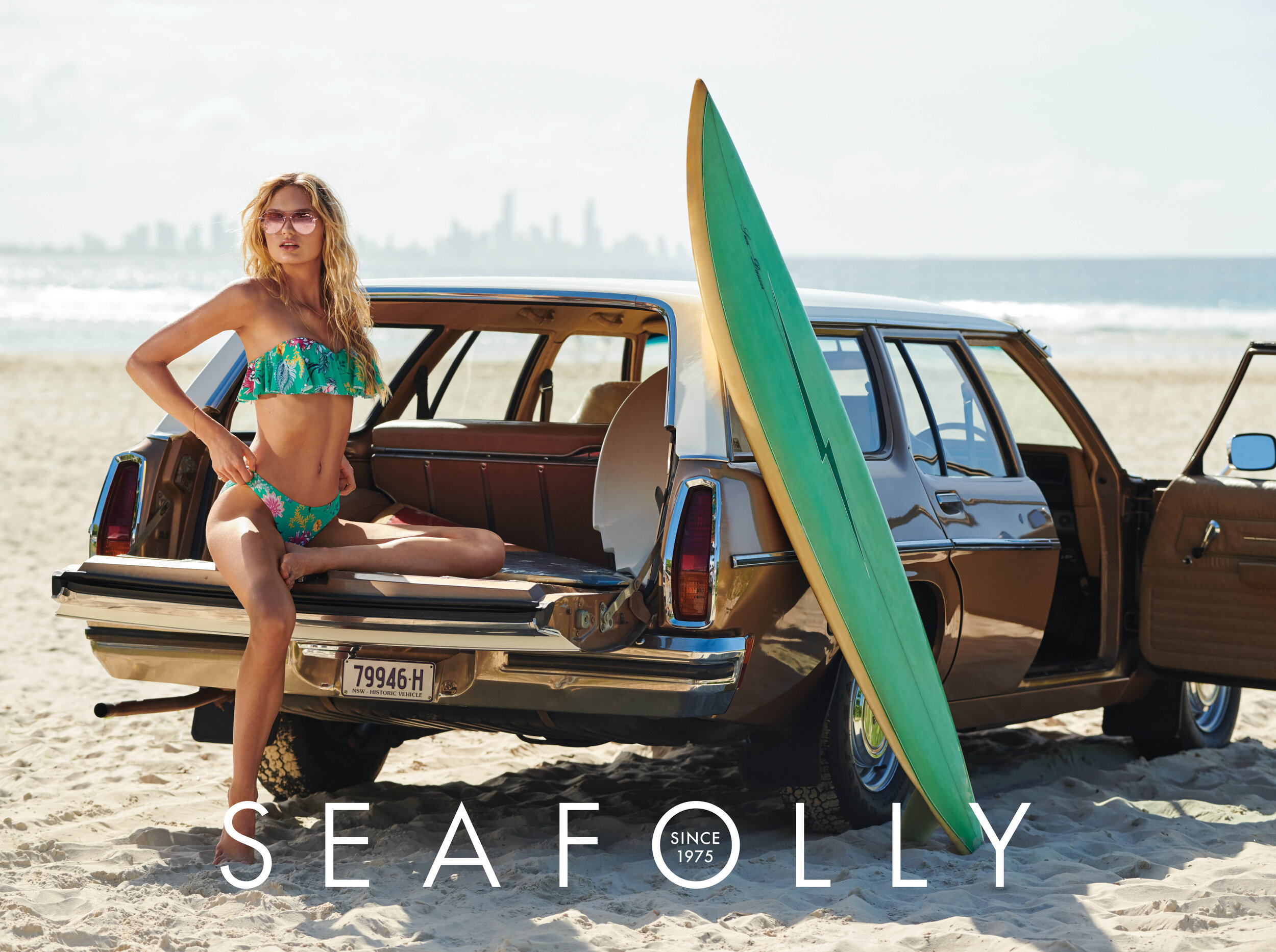 A blonde model in a floral bikini sits on the open trunk of a vintage station wagon with a surfboard on a sunny beach.