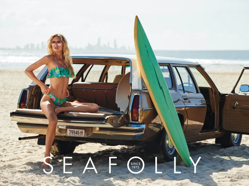 A blonde model in a floral bikini sits on the open trunk of a vintage station wagon with a surfboard on a sunny beach.