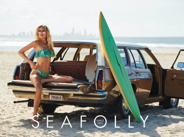 A blonde model in a floral bikini sits on the open trunk of a vintage station wagon with a surfboard on a sunny beach.