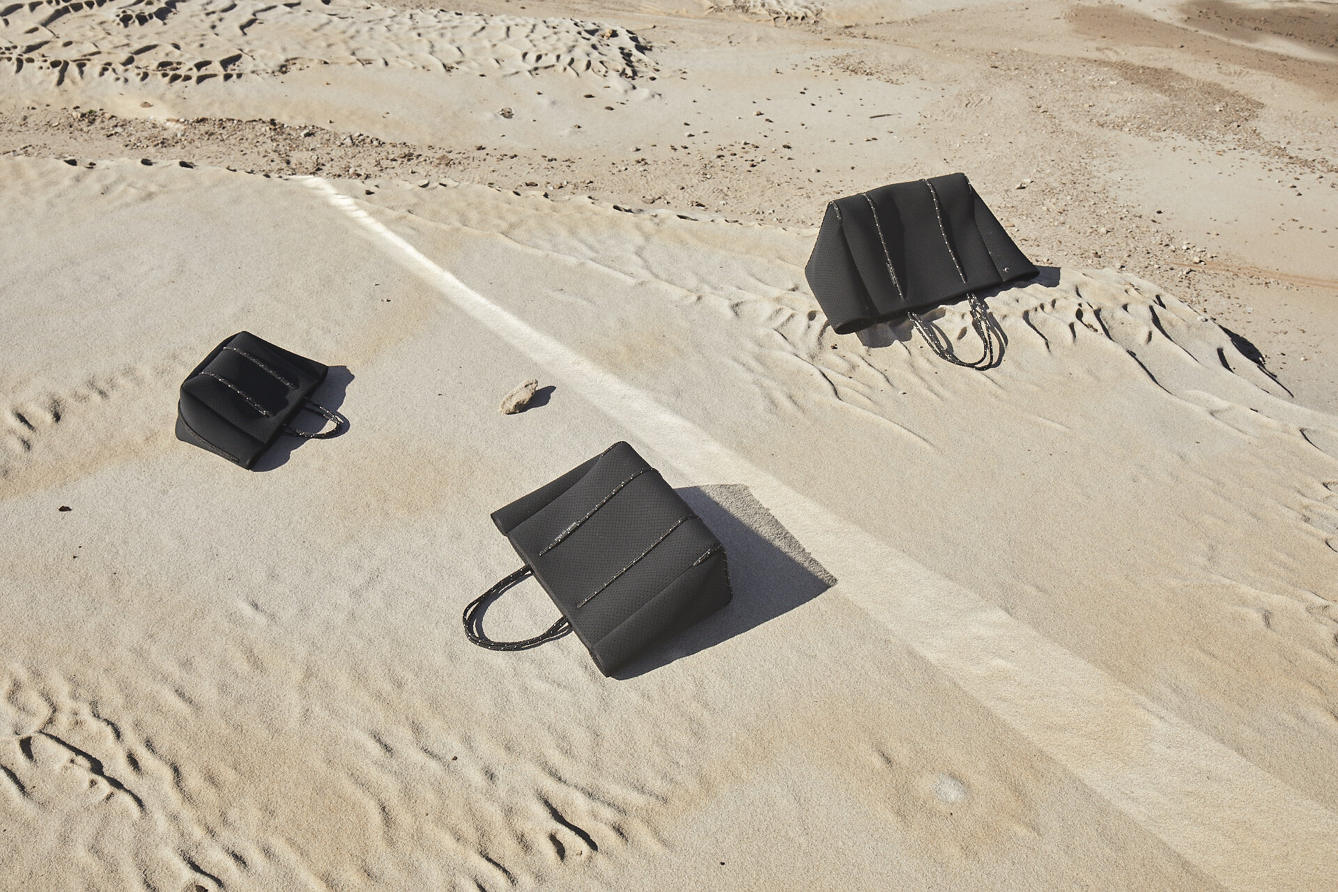 Three black tote bags are arranged on a sand dune, creating a minimalist fashion product shot in a desert environment under bright sunlight.