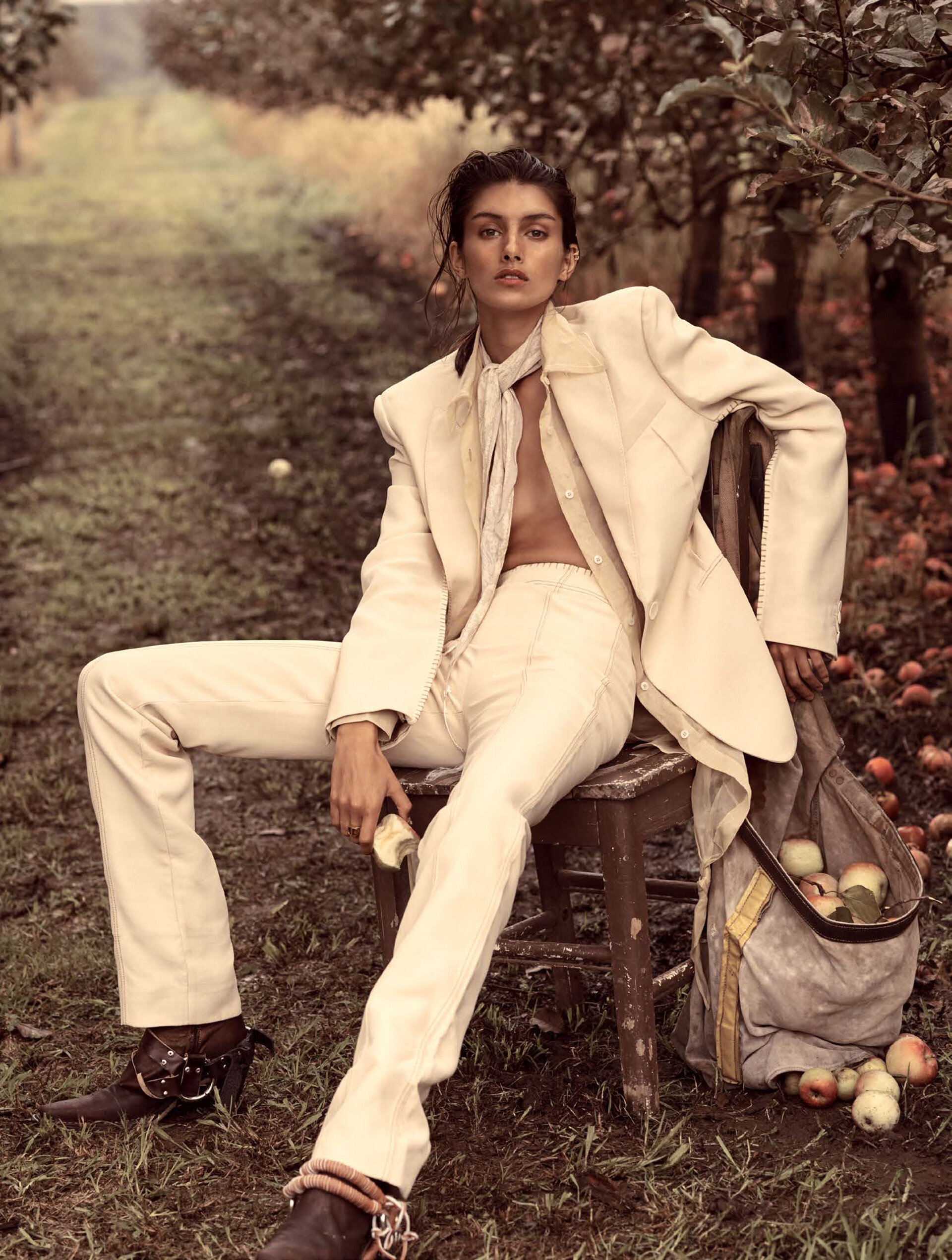 A woman in an ivory suit sits on a wooden chair in an apple orchard, surrounded by trees and apples. She holds a cut apple in her hand.