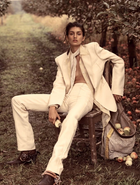 A woman in an ivory suit sits on a wooden chair in an apple orchard, surrounded by trees and apples. She holds a cut apple in her hand.