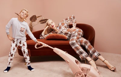 A blond woman lying on a rust-colored velvet sofa next to a young boy holding a pink vacuum cleaner in a studio setting.
