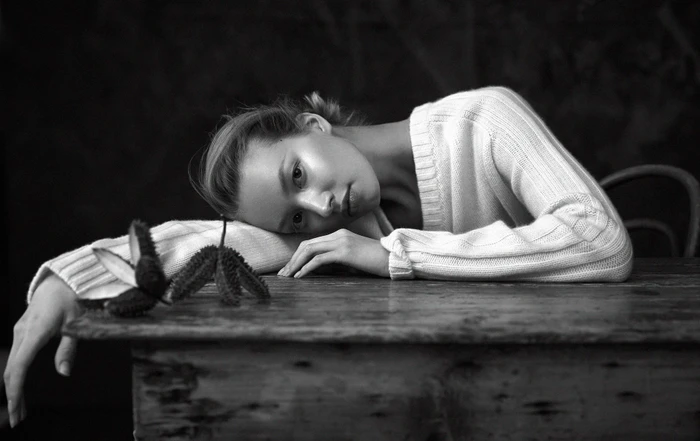 Monochrome portrait of a woman resting her head on a wooden table wearing a ribbed white sweater