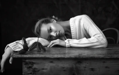 Monochrome portrait of a woman resting her head on a wooden table wearing a ribbed white sweater