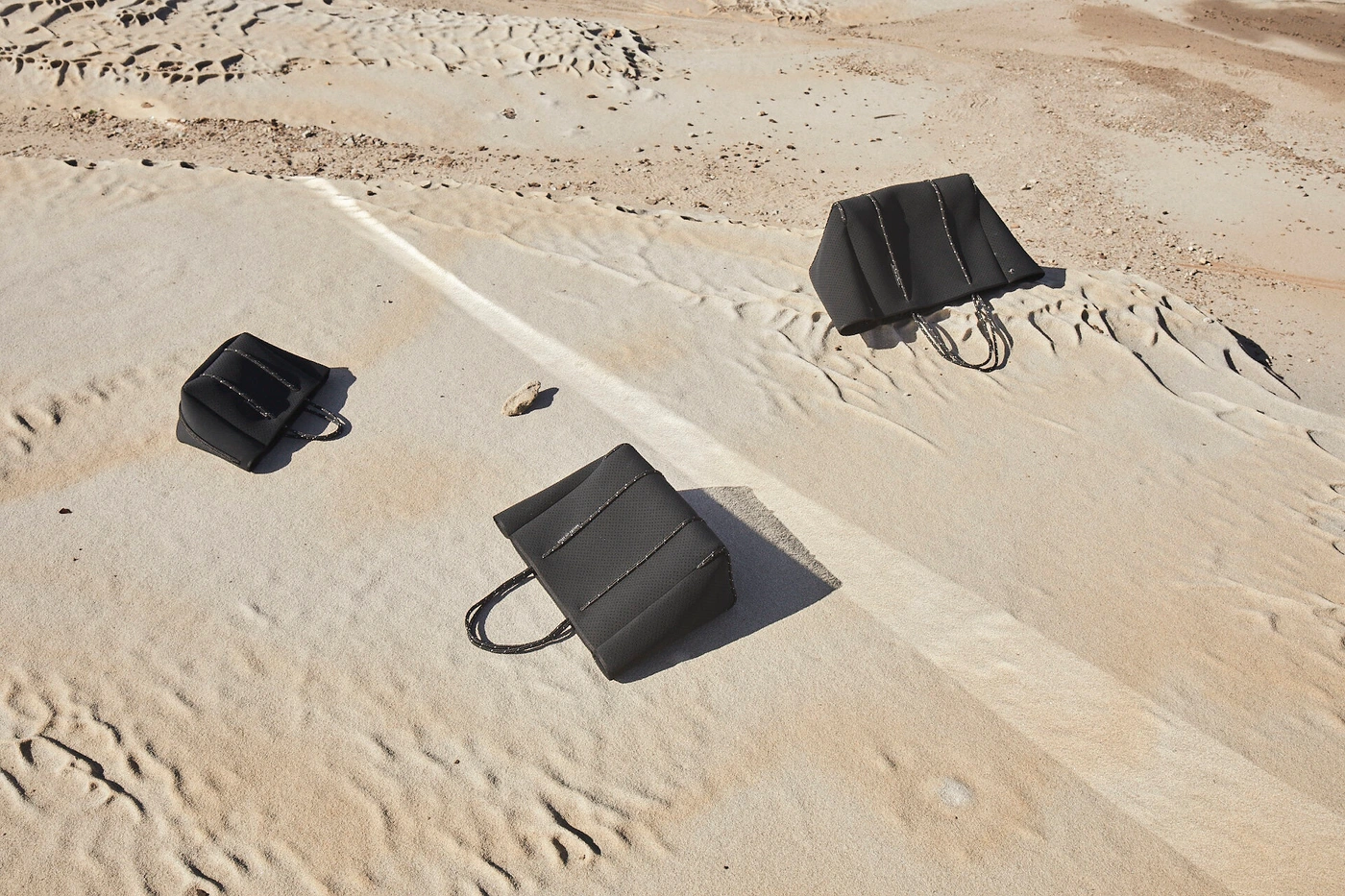 Three black handbags displayed on a textured sandy slope under bright daylight