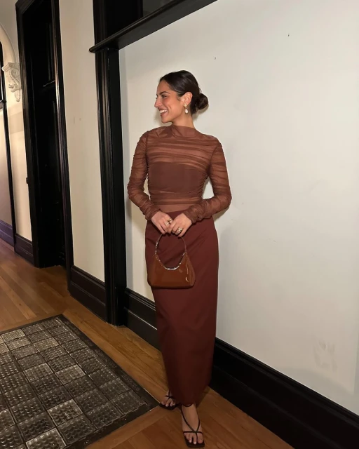 A smiling woman in a brown sheer long-sleeved top and skirt poses in a hallway with a small handbag and strappy sandals.