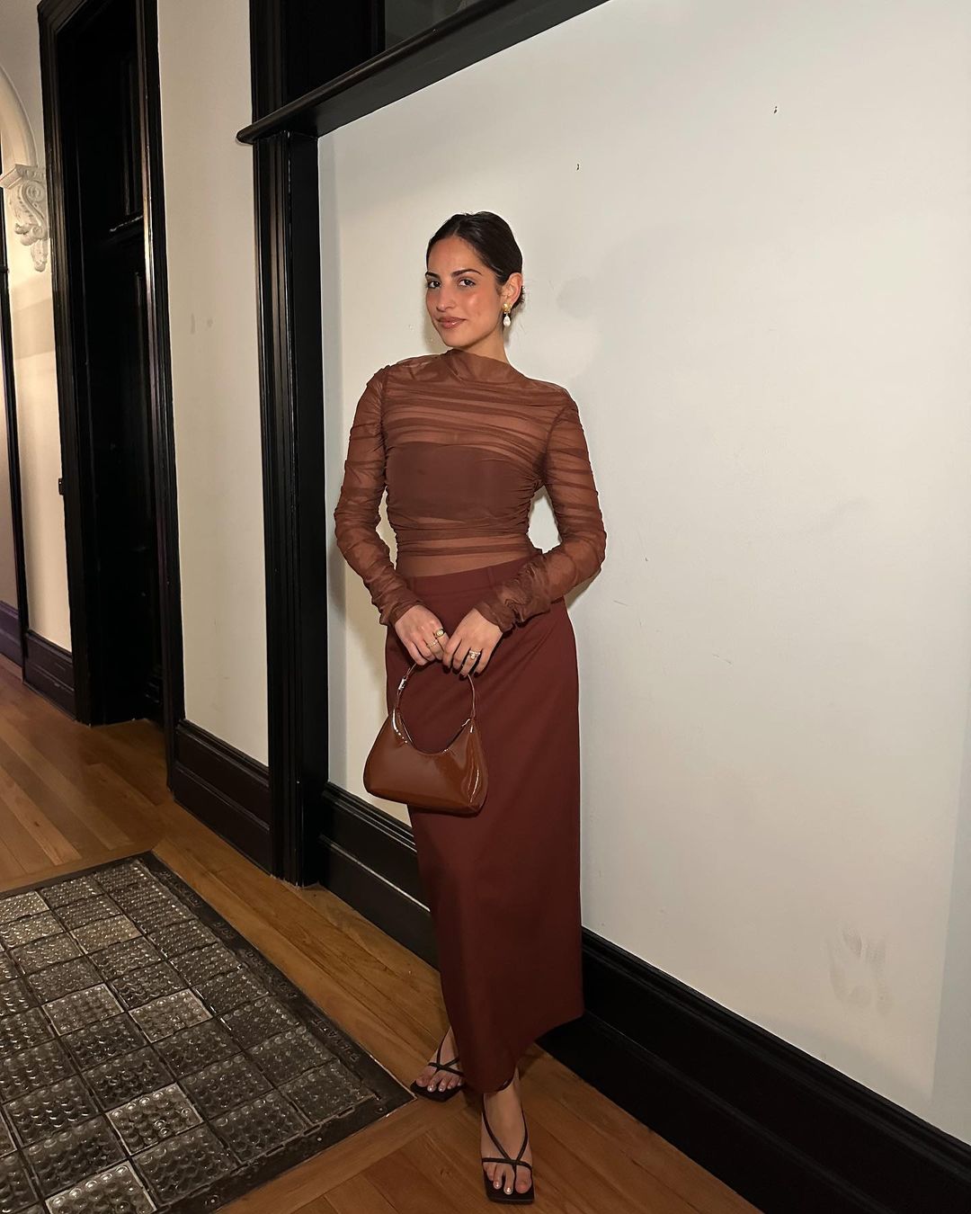 Adult woman in a brown semi-sheer top and brown skirt stands indoors holding a matching brown purse and wearing strappy sandals.