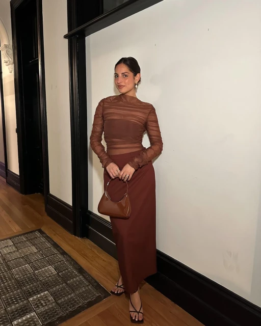 Adult woman in a brown semi-sheer top and brown skirt stands indoors holding a matching brown purse and wearing strappy sandals.