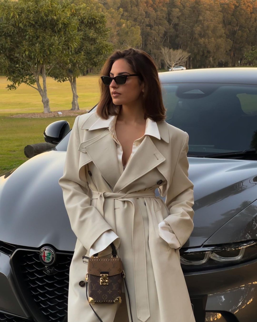 A woman in a trench coat stands next to a grey car outdoors with trees in the background. She's carrying a small patterned purse.