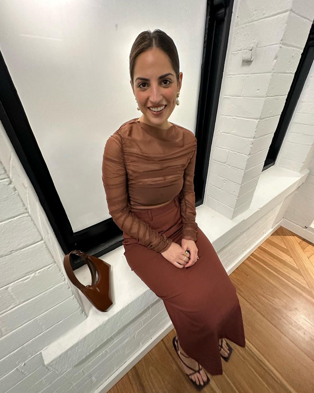 Young woman in a brown outfit poses indoors in natural light, sitting on a window ledge with a matching brown purse and strappy heels.