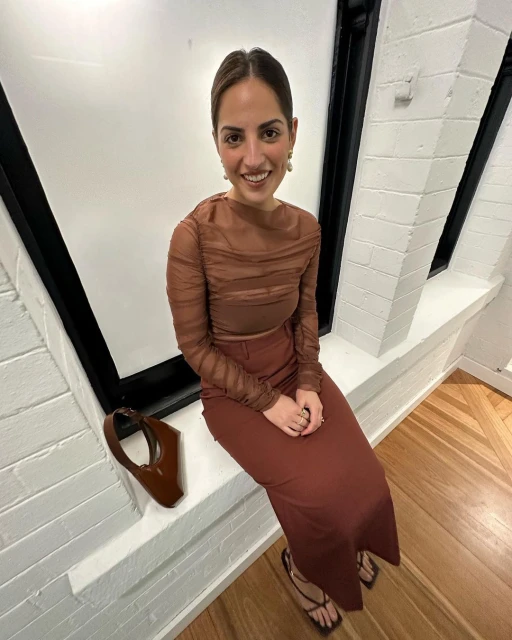 Young woman in a brown outfit poses indoors in natural light, sitting on a window ledge with a matching brown purse and strappy heels.