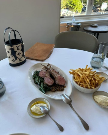Overhead shot of a restaurant table featuring lamb chops, french fries, dipping sauces, and a designer handbag, creating a luxury dining experience.