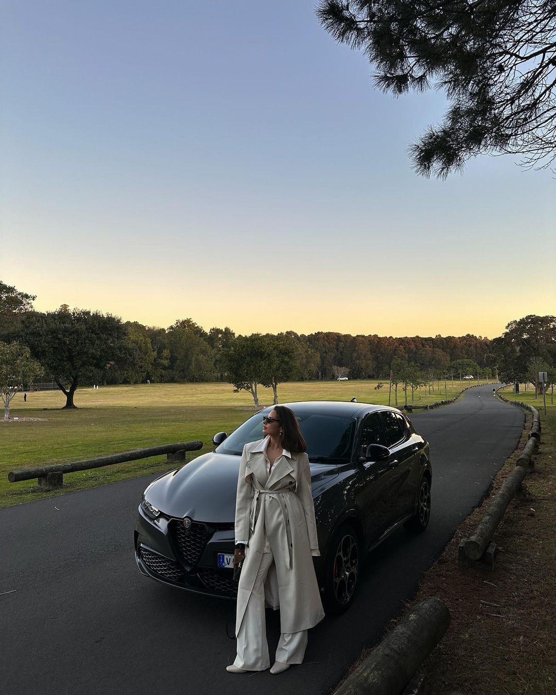 Adult female in a beige trench coat standing next to a black Alfa Romeo Tonale in a grassy area with a road at dusk.