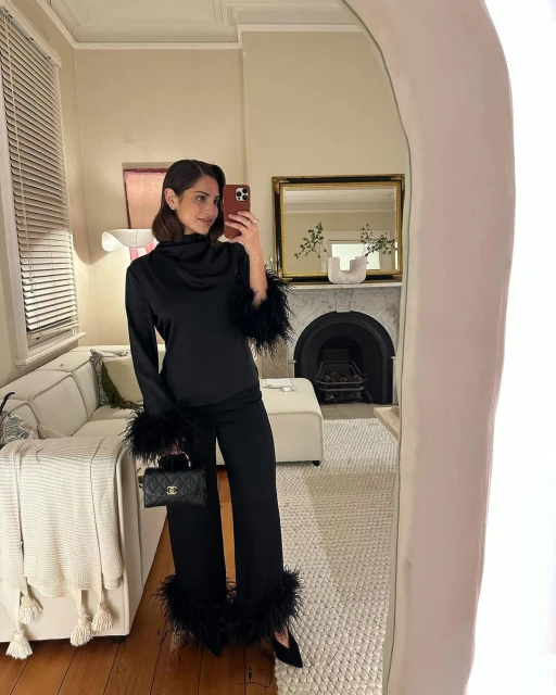 Woman takes a mirror selfie at home. She is wearing a black feather-trimmed satin pajama-style set, holding a quilted handbag.