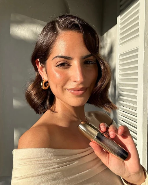 Close-up of a woman with brunette wavy hair and gold hoop earrings, holding a gold and black cosmetic tube in her right hand.