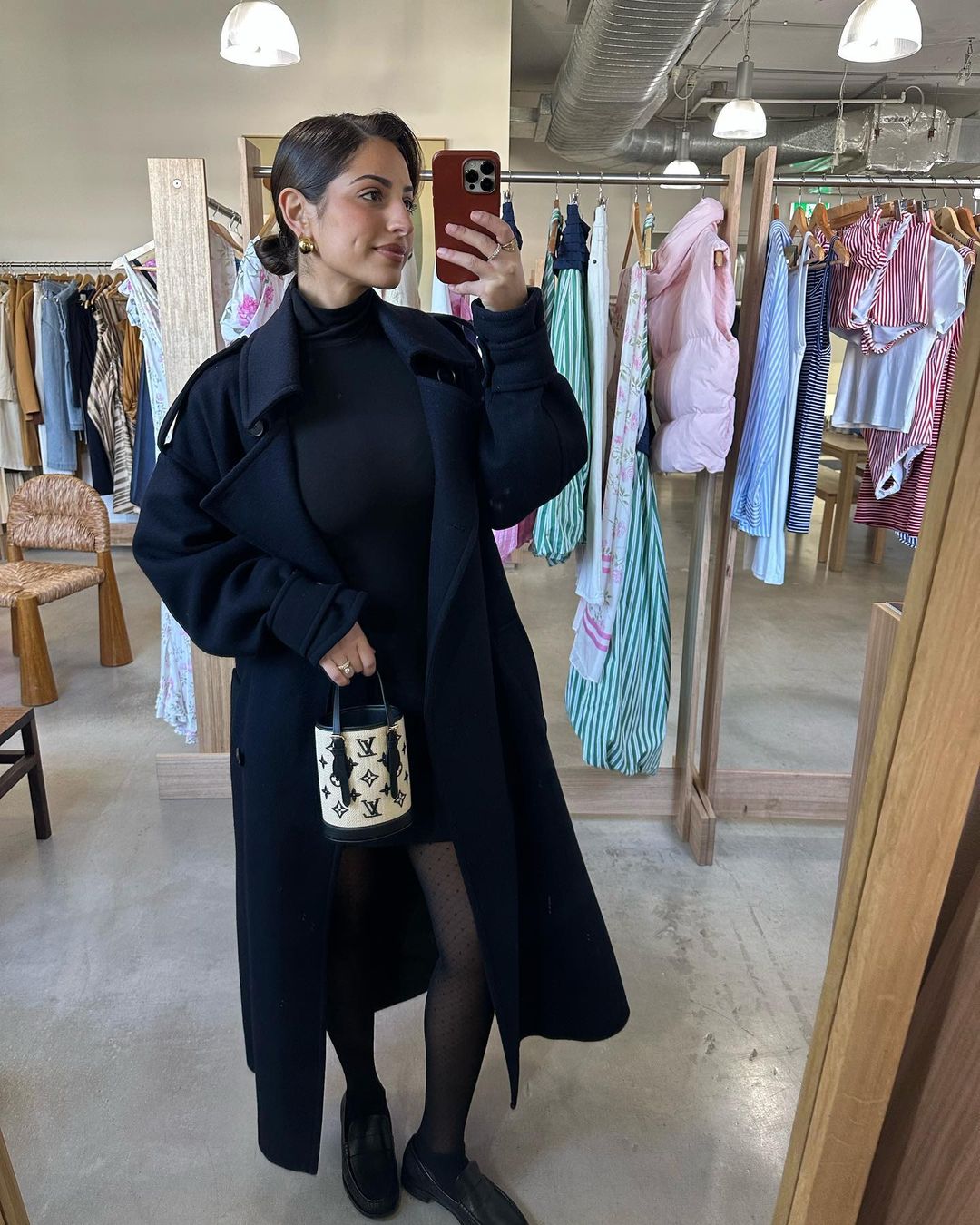 Woman in black coat holds handbag in clothing store, posing in mirror selfie for social media, showcasing fashion and style.