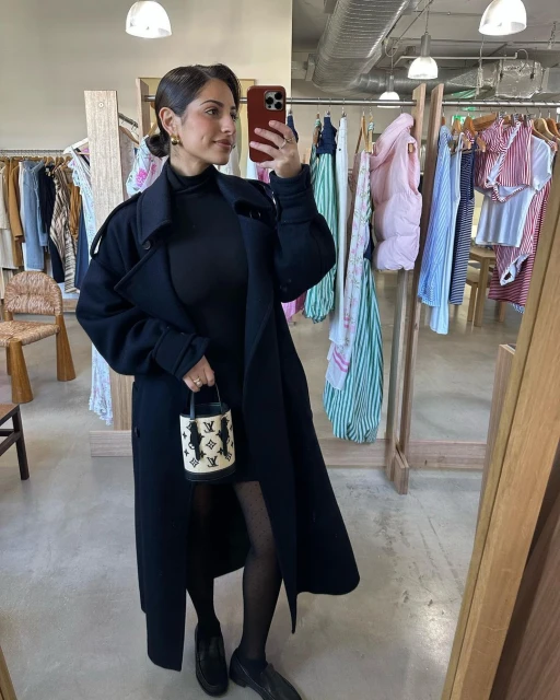 Woman in black coat holds handbag in clothing store, posing in mirror selfie for social media, showcasing fashion and style.
