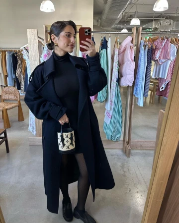 Woman in black coat holds handbag in clothing store, posing in mirror selfie for social media, showcasing fashion and style.