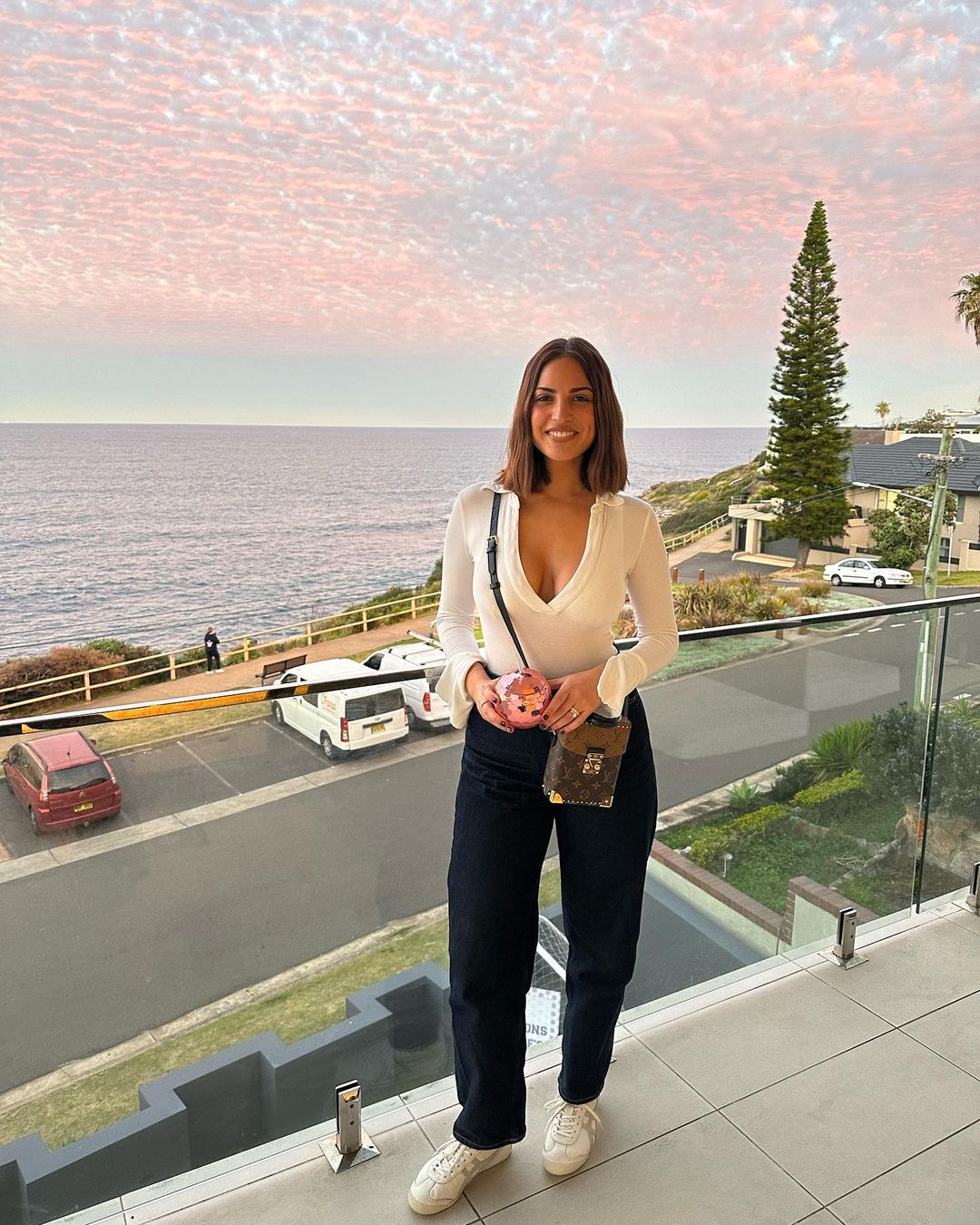 A woman in a white top and dark jeans stands on a balcony overlooking the ocean and sky at dusk wearing designer bags.