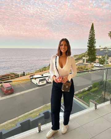 A woman in a white top and dark jeans stands on a balcony overlooking the ocean and sky at dusk wearing designer bags.